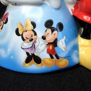 May include: A ceramic figurine of Mickey Mouse with a blue and white background. Mickey is wearing red shorts and yellow shoes. Minnie Mouse is wearing a purple dress and yellow shoes. The figurine is decorated with white glitter.