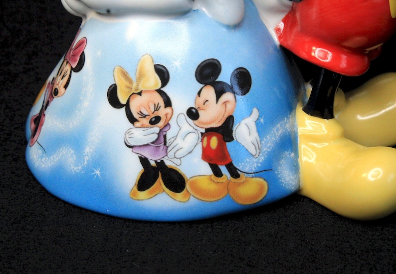 May include: A ceramic figurine of Mickey Mouse with a blue and white background. Mickey is wearing red shorts and yellow shoes. Minnie Mouse is wearing a purple dress and yellow shoes. The figurine is decorated with white glitter.