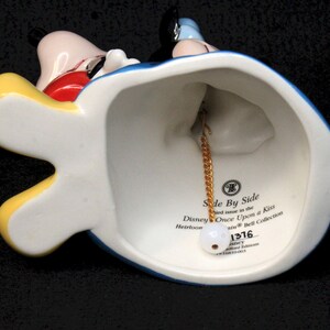 May include: A porcelain bell featuring Mickey Mouse and Minnie Mouse in their iconic outfits. The bell is blue, yellow, and red with a gold chain and a white tag that reads "Side By Side and issue in the Disney Once Upon a Kiss Heirloom Bell Collection 1376".