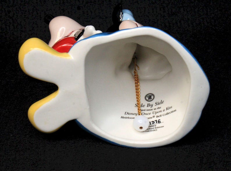May include: A porcelain bell featuring Mickey Mouse and Minnie Mouse in their iconic outfits. The bell is blue, yellow, and red with a gold chain and a white tag that reads "Side By Side and issue in the Disney Once Upon a Kiss Heirloom Bell Collection 1376".