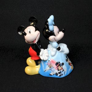 May include: A ceramic figurine of Mickey Mouse and Minnie Mouse. Mickey is wearing his signature red shorts and yellow shoes. Minnie is wearing a blue dress with a white bow. The figurine is decorated with images of other Disney characters.