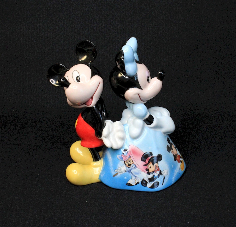 May include: A ceramic figurine of Mickey Mouse and Minnie Mouse. Mickey is wearing his signature red shorts and yellow shoes. Minnie is wearing a blue dress with a white bow. The figurine is decorated with images of other Disney characters.