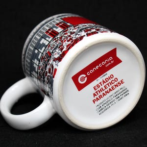 May include: White ceramic mug featuring a stadium design in red, gray, and black. The mug has a curved handle and a red label with text. The bottom of the mug has the text "EST&Aacute;DIO ATHLETICO PARANAENSE".