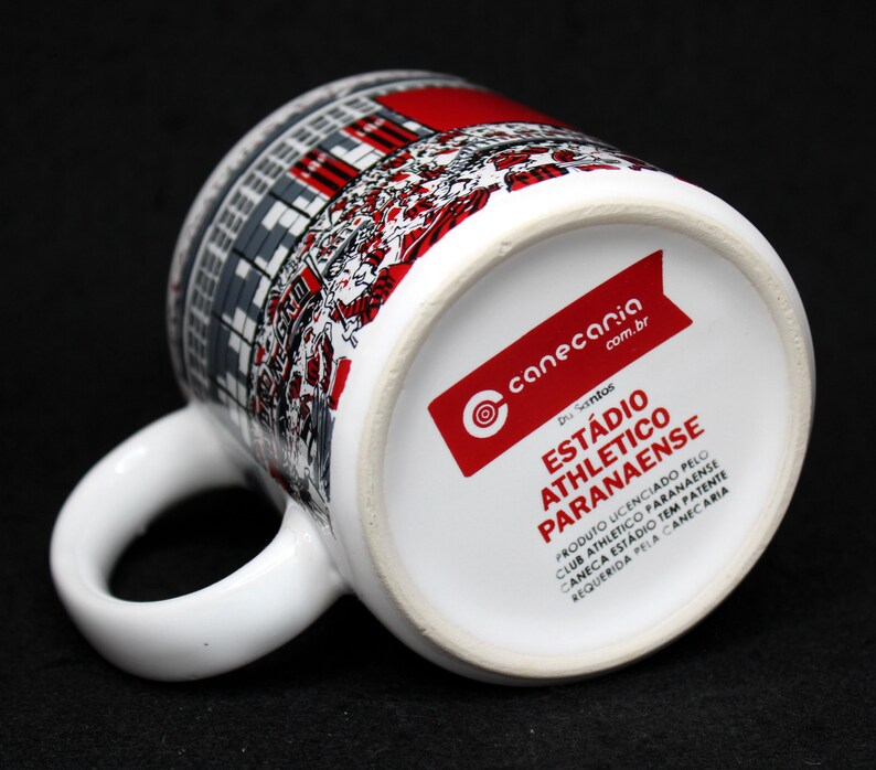 May include: White ceramic mug featuring a stadium design in red, gray, and black. The mug has a curved handle and a red label with text. The bottom of the mug has the text "EST&Aacute;DIO ATHLETICO PARANAENSE".