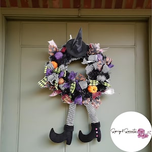 Puede incluir: Una corona de Halloween con un sombrero de bruja negro, piernas a rayas blancas y negras y zapatos negros. La corona está decorada con detalles morados, naranjas y negros, incluyendo lazos y calabazas. Cuelga de una puerta verde.