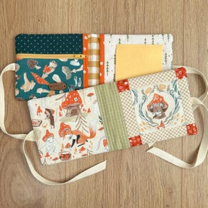 May include: A fabric book with mushroom and forest animal designs. It has a zipped pocket, a yellow felt pocket, and panels of green, orange, and beige patterned fabric. Cream straps are attached for carrying. The book is handmade.