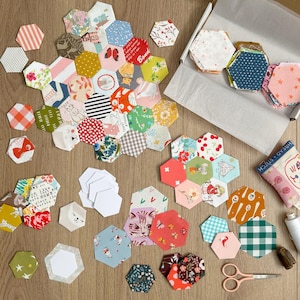 May include: A collection of colourful fabric hexagons, some with patterns and some with images, are arranged on a wooden surface. There are also several white paper hexagons and a pair of pink scissors.
