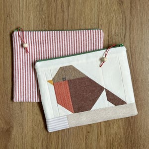 以下が含まれることがあります： 2つの布製ポーチ。1つは白地に茶色、オレンジ、黄色の幾何学的な鳥のデザイン。もう1つは赤と白のストライプ。どちらも緑色のジッパーと赤い紐に木製のビーズが付いています。