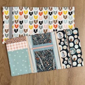 May include: Two fabric organizers. The top one has a white background with repeating cat faces in various colors. The bottom organizer has multiple patterned fabrics, including floral, gingham, and a dark blue with forest animals. It features clear pockets and zippers.