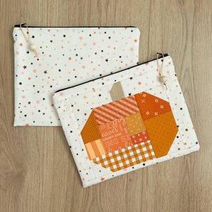 May include: Two rectangular zippered pouches made of white fabric with a pattern of small black and orange stars. One pouch features a quilted pumpkin design composed of orange and white fabric patches with plaid and text patterns.