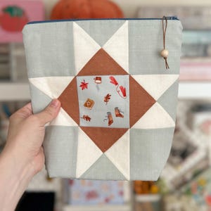 May include: A fabric pouch with a star pattern in pale blue, white, and brown. The central square features illustrations of autumn leaves, a mug, mittens, and boots. A blue zip and a wooden bead accent the top.