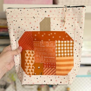 May include: A zippered pouch made of cream fabric with a pink and brown star pattern. The front features a patchwork pumpkin design in shades of orange, brown, and white, with text including "chilly," "pumpkin," and "apple picking."