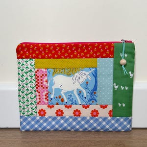 May include: A patchwork fabric pouch with a unicorn illustration. The design incorporates red, green, yellow, and pink patterned fabrics. The pouch has a pink zip and a green tassel with a wooden bead. The overall design is a mix of patterns and colours.