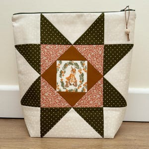 May include: A zipped pouch featuring a patchwork star design in olive green, cream, and coral floral fabric. The centre square displays a printed illustration of rabbits surrounded by a wreath of orange berries and green leaves, with a wooden bead pull.