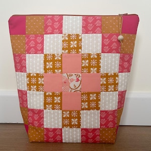 May include: A patchwork fabric tote bag in shades of pink, peach, white, and gold. The bag features a zip closure and a wooden bead on the zip pull. The design is a grid of squares with floral and geometric patterns.