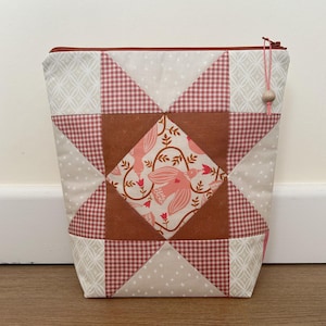May include: A patchwork fabric pouch with a star design. The bag features cream, red, and brown squares and triangles. The central square has a bird and floral pattern. A red zip and a wooden bead adorn the top.