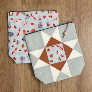 May include: Two fabric pouches with zip closures. One is light blue with a repeating pattern of autumn-themed illustrations. The other has a geometric star design in shades of grey, white, and brown, with a fall-themed centre.