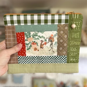 May include: A patchwork zipped pouch with a garden scene illustration of a frog and mice harvesting radishes. The pouch has a green and white gingham top, a brown and white patterned side, and a green zip. A wooden bead hangs from the zip pull.