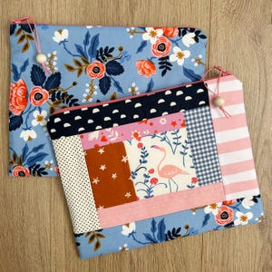 May include: Two rectangular zippered pouches. One features a light blue fabric with a floral pattern of orange and white flowers. The other is a patchwork design with a pink flamingo, stars, polka dots, and stripes, finished with a wooden bead pull.