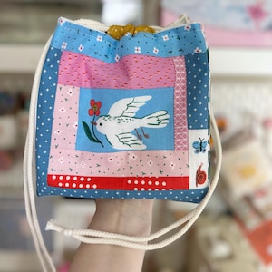 May include: A patchwork drawstring bag with a bird and flower design. The bag features a blue, pink, and red color scheme with white floral and polka dot patterns. A white bird with a flower is centered on the front. The bag has a white drawstring closure.