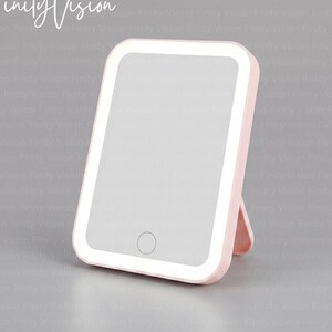 May include: A pink, rectangular vanity mirror with a built-in LED light and a stand. The mirror has a touch-sensitive button and the text "Finity Vision" in the upper left corner.