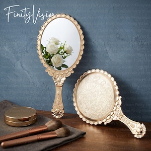 May include: Two gold-toned hand mirrors with ornate scalloped edges and intricate embossed handles. One mirror reflects white roses, while the other displays a detailed floral relief pattern on its back. Two makeup brushes and a compact are present.