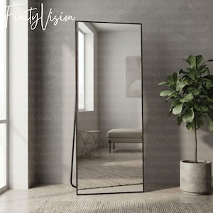 May include: A tall, rectangular floor mirror with a thin, dark metal frame. The mirror is positioned at a slight angle, reflecting a room interior that includes a light-colored bench, a small geometric side table, and a window with natural light.