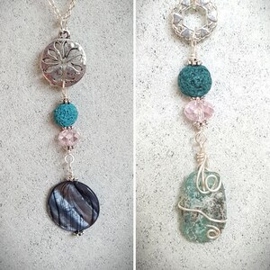 May include: Two silver necklaces with different pendants. The necklace on the left has a silver circle pendant with a floral design, a teal lava bead, a pink crystal bead, and a black and silver shell pendant. The necklace on the right has a silver pendant with a circle of hearts, a teal lava bead, a pink crystal bead, and a teal and silver wrapped glass pendant.