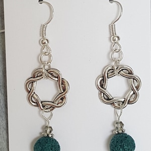 May include: Silver earrings with a knotted circle design and a dark green lava rock bead. The earrings are on a white card with the text "Surely Jane's Designs".