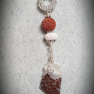 May include: A silver chain necklace with a silver heart-shaped ring, a red lava rock bead, a white bead with a sparkly finish, and a brown and white agate pendant.
