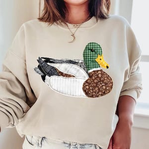 Op de afbeelding: Beige sweatshirt met een applicatie van een eend. De eend heeft een groen geruite kop, een bruin bloemenlichaam en een gele snavel. De vleugels van de eend zijn een mix van grijze, witte en bruine stoffen. De sweatshirt heeft een ronde hals en lange mouwen.