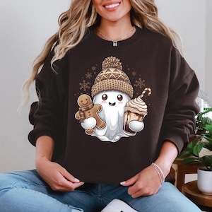 Może przedstawiać: Ciemnobrązowa bluza z kapturem z kreskówkowym duchem w dzianinowej czapce trzymającym piernikowego ludzika i gorącą czekoladę. Duch jest biały z uśmiechniętą twarzą. Czapka jest brązowo-beżowa z pomponem. Płatki śniegu w tle.