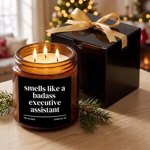Puede incluir: Una vela de tres mechas en un frasco de vidrio ámbar con una etiqueta negra que dice "smells like a badass executive assistant" y "net weight 16oz / 454g". Junto a ella, una caja de regalo negra con un lazo dorado y ramas de pino.