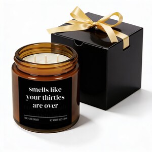 Puede incluir: Una vela en tarro de cristal marr&oacute;n con tres mechas y una etiqueta negra que dice "Smells like your thirties are over". La vela est&aacute; junto a una caja de regalo negra atada con una cinta dorada. La vela pesa 510g.