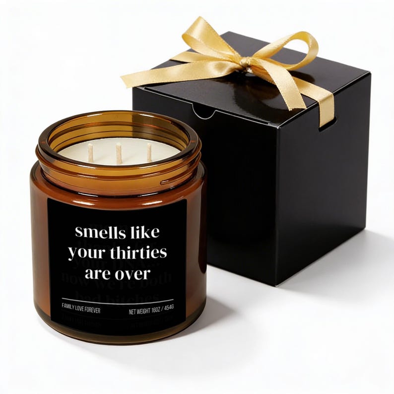 Puede incluir: Una vela en tarro de cristal marr&oacute;n con tres mechas y una etiqueta negra que dice "Smells like your thirties are over". La vela est&aacute; junto a una caja de regalo negra atada con una cinta dorada. La vela pesa 510g.