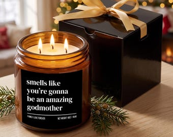 May include: A three-wick candle in an amber glass jar with a black label reading "smells like you're gonna be an amazing godmother" and "NET WEIGHT 16OZ / 454G". Beside it sits a black gift box topped with a gold ribbon and green pine sprigs.