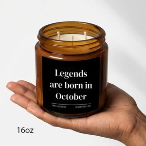 May include: A brown glass jar candle with three wicks. The black label reads "Legends are born in October." The candle is held in a hand. The bottom left corner of the image shows "16oz".