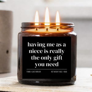 Puede incluir: Una vela encendida en un tarro de cristal marrón con una etiqueta negra. La etiqueta dice "having me as a niece is really the only gift you need". La vela tiene tres mechas y el texto "FAMILY LOVE FOREVER" y "NET WEIGHT 16OZ / 454G".