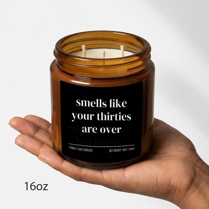 Puede incluir: Una vela en tarro de cristal marr&oacute;n con una etiqueta negra que dice "smells like your thirties are over". La vela tiene tres mechas y se sostiene en una mano. El peso neto es de 454g.