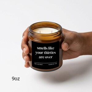 Puede incluir: Un tarro de vela de vidrio marr&oacute;n con una etiqueta negra que dice "smells like your thirties are over". La vela tiene dos mechas y una cera blanca. El tarro se sostiene en una mano. El texto "9oz" est&aacute; en la parte inferior.
