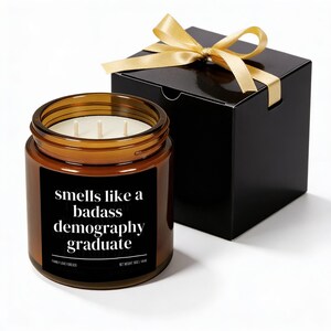 May include: A brown glass jar candle with three wicks and a black label that reads "smells like a badass demography graduate." The candle is next to a black gift box tied with a gold ribbon.