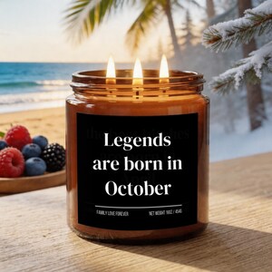 May include: A lit candle in a glass jar with a black label that reads "Legends are born in October." The candle has three wicks and is surrounded by a blurred background of a beach scene and a wooden bowl of berries.