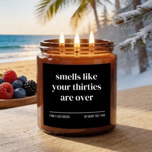 Puede incluir: Una vela encendida de tres mechas en un tarro de cristal marr&oacute;n con una etiqueta negra. La etiqueta dice "smells like your thirties are over". La vela es de 454g. Hay bayas en un cuenco de madera en el fondo. La vela est&aacute; sobre una superficie de madera.