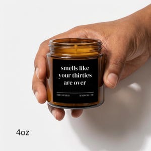 Puede incluir: Una vela en tarro de cristal marr&oacute;n con una etiqueta negra que dice "smells like your thirties are over". La vela se sostiene en una mano. El peso neto es de 113g.