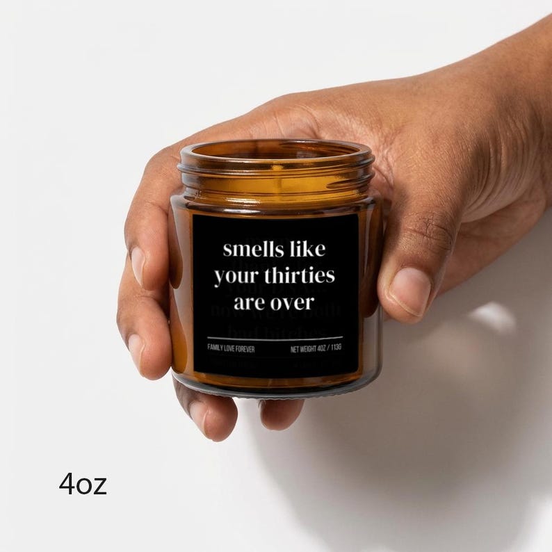 Puede incluir: Una vela en tarro de cristal marr&oacute;n con una etiqueta negra que dice "smells like your thirties are over". La vela se sostiene en una mano. El peso neto es de 113g.
