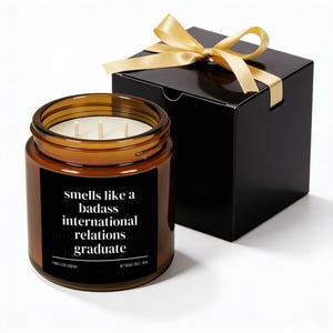May include: A brown glass jar candle with three wicks and a black label that reads "smells like a badass international relations graduate." The candle is next to a black gift box tied with a gold ribbon.