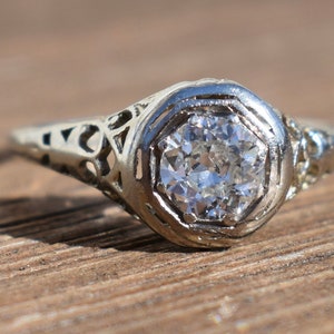 May include: A vintage-style engagement ring with a round, clear diamond set in a white metal filigree setting. The ring's band features intricate openwork designs. The ring is set against a wooden surface.