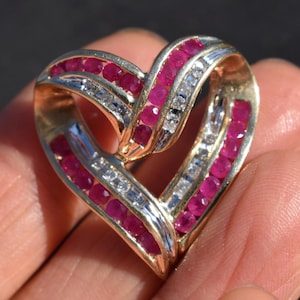 May include: A heart-shaped pendant with a gold frame, encrusted with vibrant red rubies and sparkling white diamonds. The pendant's design features a layered, open-heart shape, showcasing the gemstones' brilliance. A beautiful piece of jewelry.
