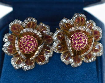 May include: A pair of floral earrings with a gold and red enamel design. The earrings feature a central cluster of small red gemstones surrounded by petals with diamond accents. The earrings are displayed in a blue jewelry box.
