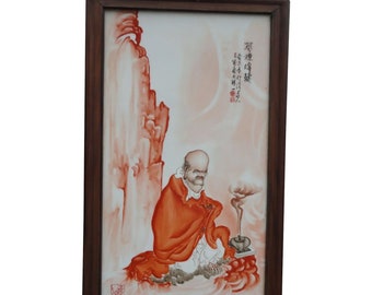 May include: A framed porcelain tile with a painting of a man in a red robe meditating in front of a burning incense stick. The tile has a light pink background with a mountain in the background. The tile is framed in a dark wood frame.